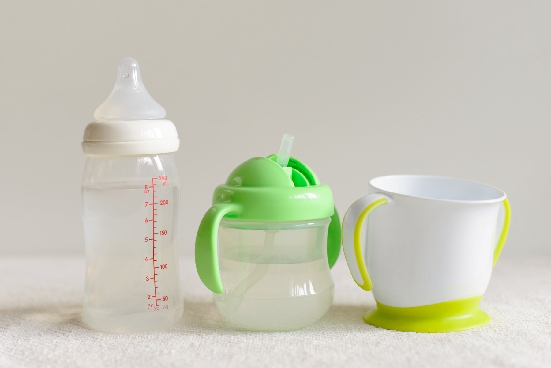 A white background and soft towel like surface. From left to right, a clear baby bottle, a sippy cup with a green lid, and a plastic cup designed for babies and toddlers
