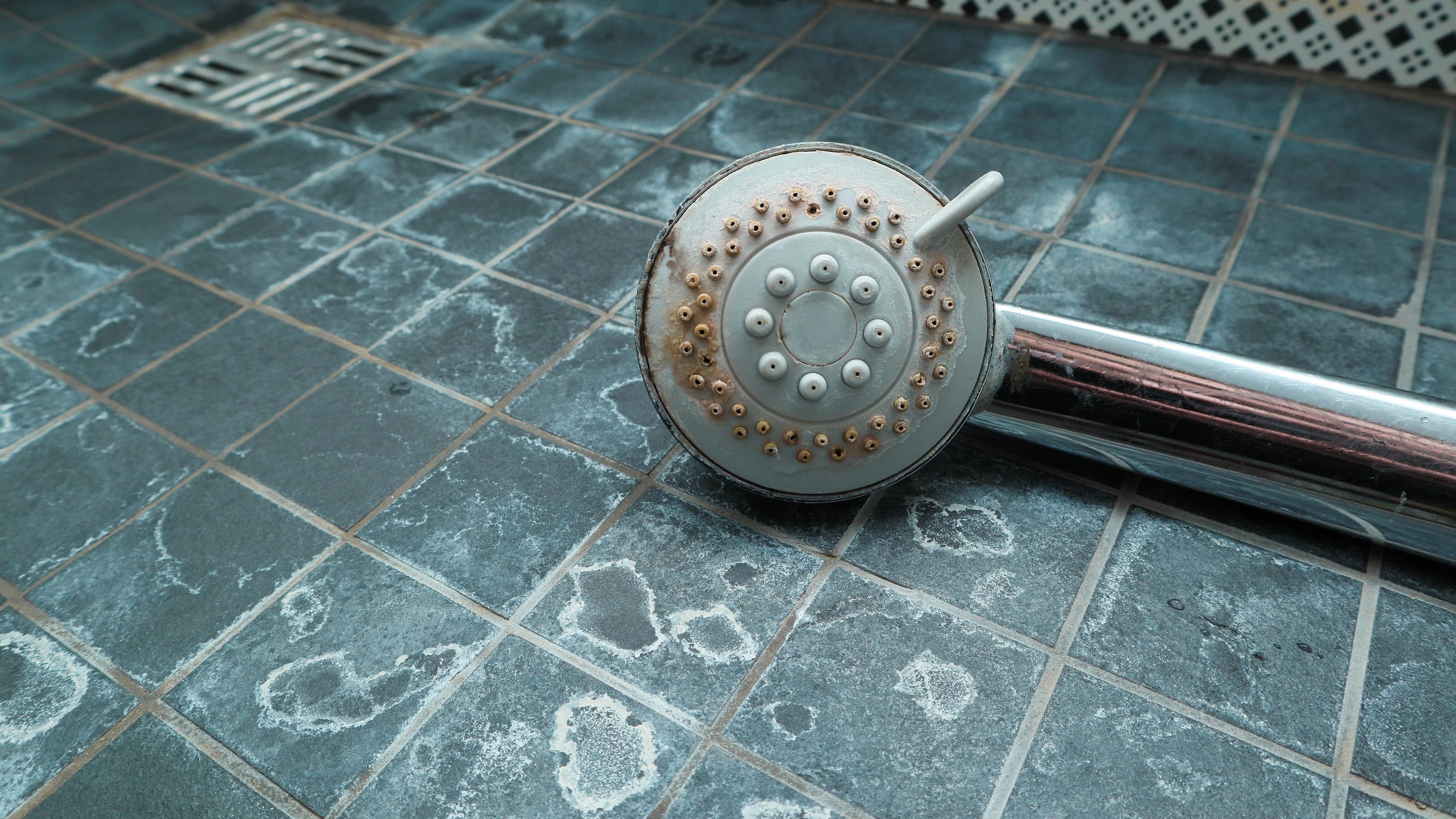 Dirty chrome shower head on a shower tile floor, both with hard water scale buildup