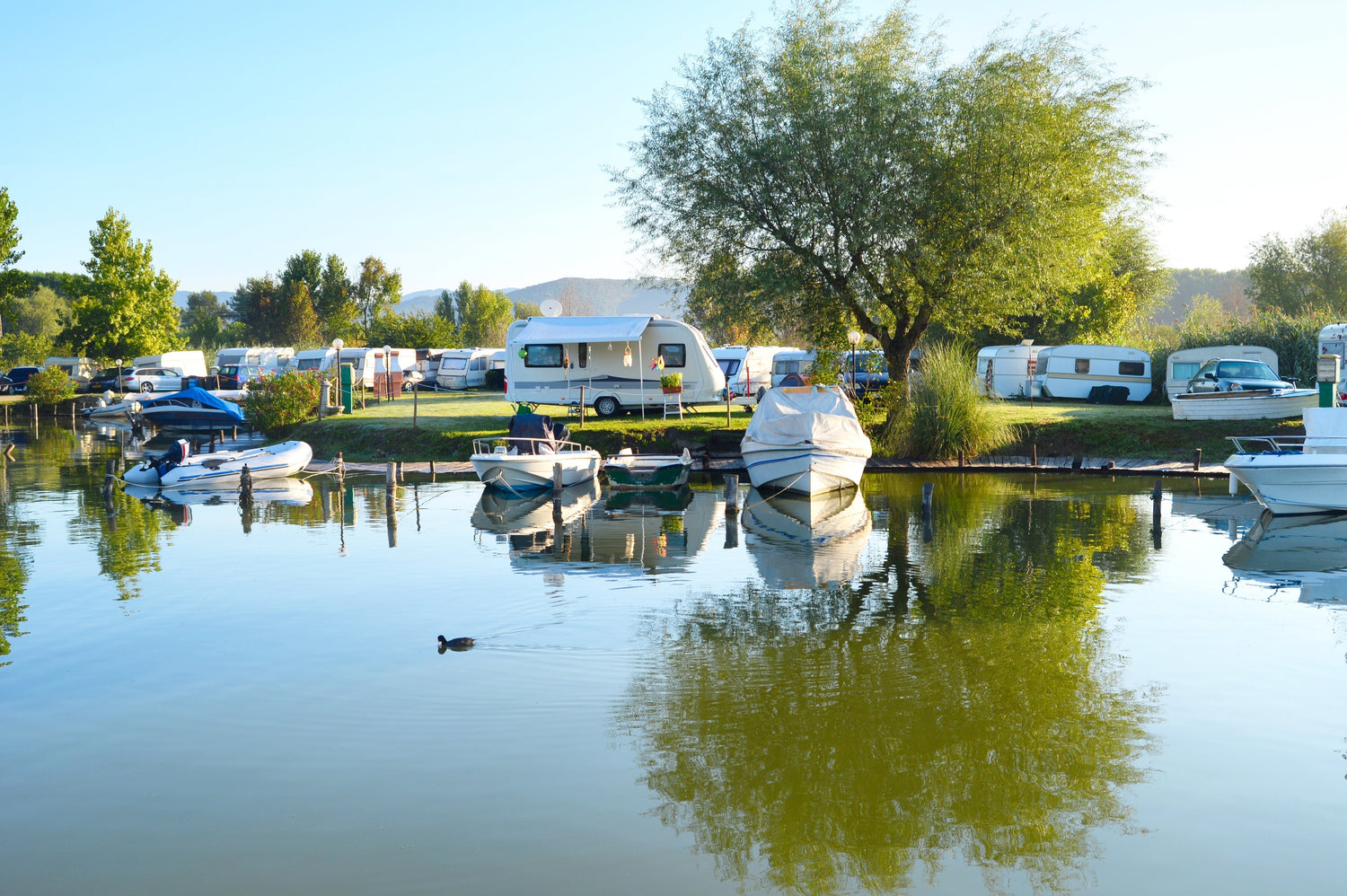 A campsite situated next to a freshwater lake with green lawn and trees. Rvs, campers, and docked boats are within frame